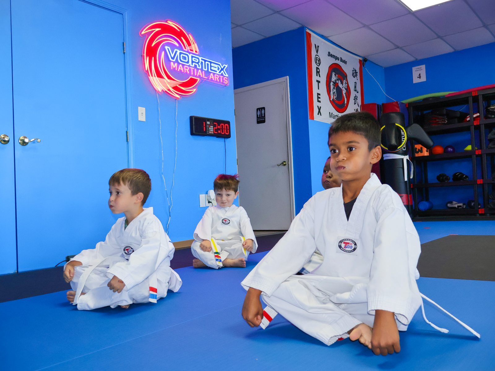 Young students in white gis sitting on the mat in the Vortex training room, with the academy neon sign on the blue wall behind them