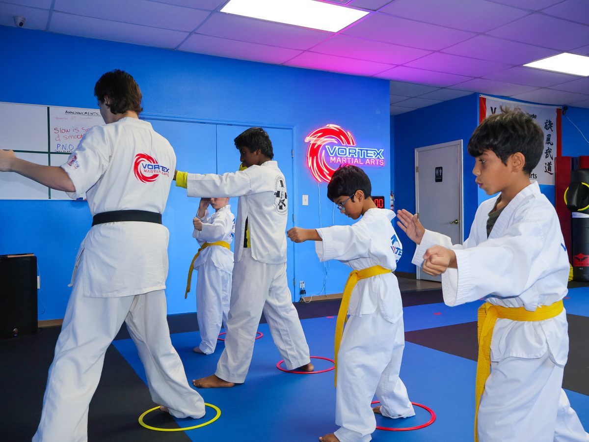 Kids in white gis with gold belts throwing punches in a line while an instructor leads the drill