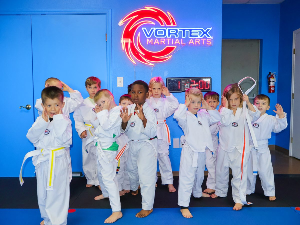A group of preschoolers in white gis holding their hands up in a guard stance under the Vortex Martial Arts sign