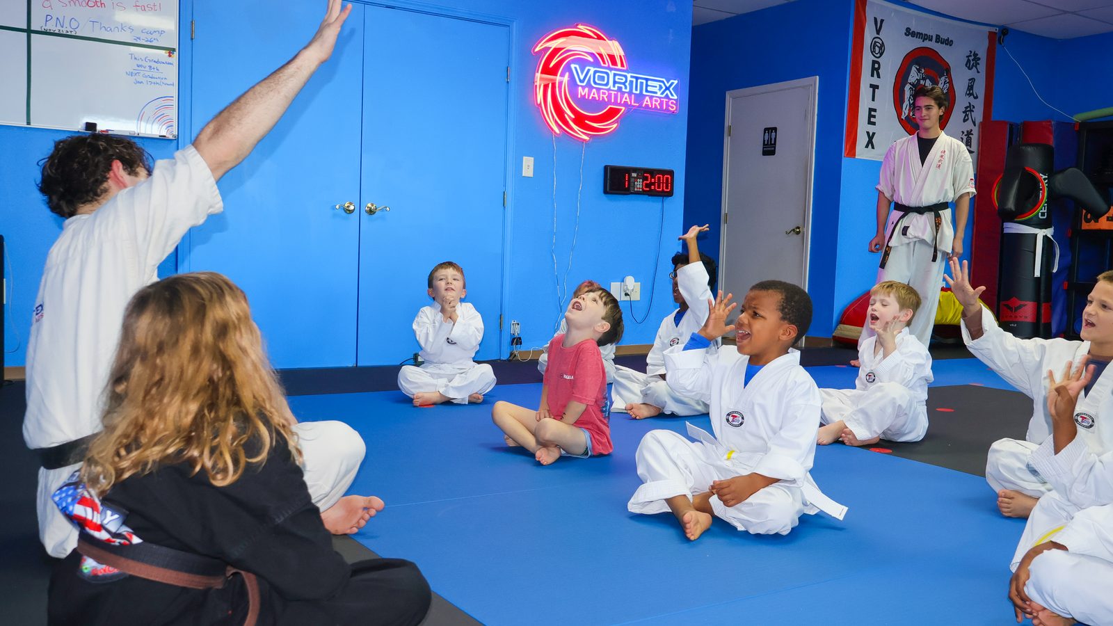 A big group of kids sitting on the mat with hands raised, laughing during a class game