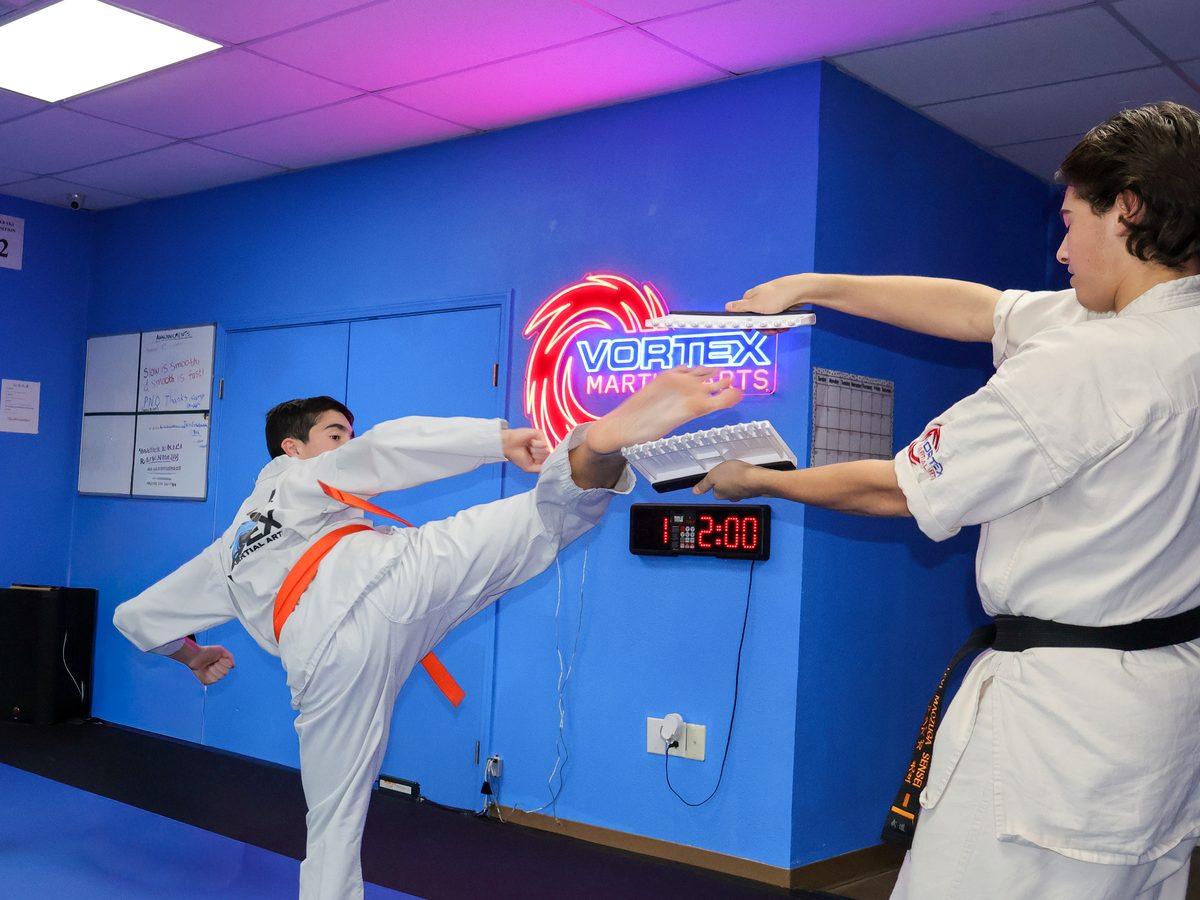A teen student in a white gi with an orange belt breaking a board with a jumping kick while a black belt holds it